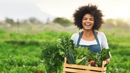 Mujer sonriente con un delantal y una caja de madera llena de productos frescos caminando fuera de un campo.